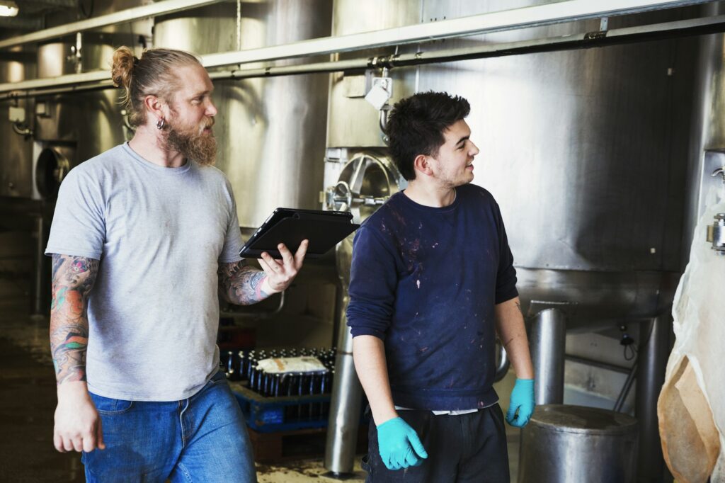 Two men working in a brewery, walking past large metal kettles, holding digital tablet.